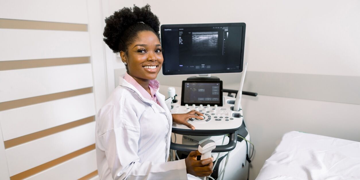Smiling African woman doctor with ultrasound scanner in hand, working on modern ultrasound scanning machine in light room in clinic. Portrait Of 4D Ultrasound Scanning Machine Operator